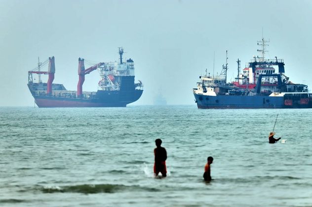 Dos niños se encuentran en aguas poco profundas con flotadores de espuma mientras se ven barcos de carga y otras embarcaciones en el Estrecho de Ormuz, frente a Bandar Abbas, Irán, el 30 de junio de 2026. (Amirhosein Khorgooi)