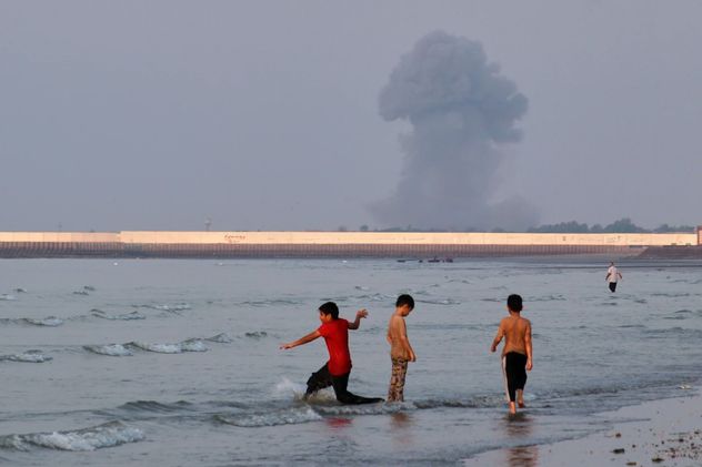 Tres niños juegan en las aguas poco profundas del Estrecho de Ormuz, mientras una columna de humo se eleva de una explosión de fondo, frente a Bandar Abbas, Irán, el 13 de julio de 2026. (Razieh Poudat)