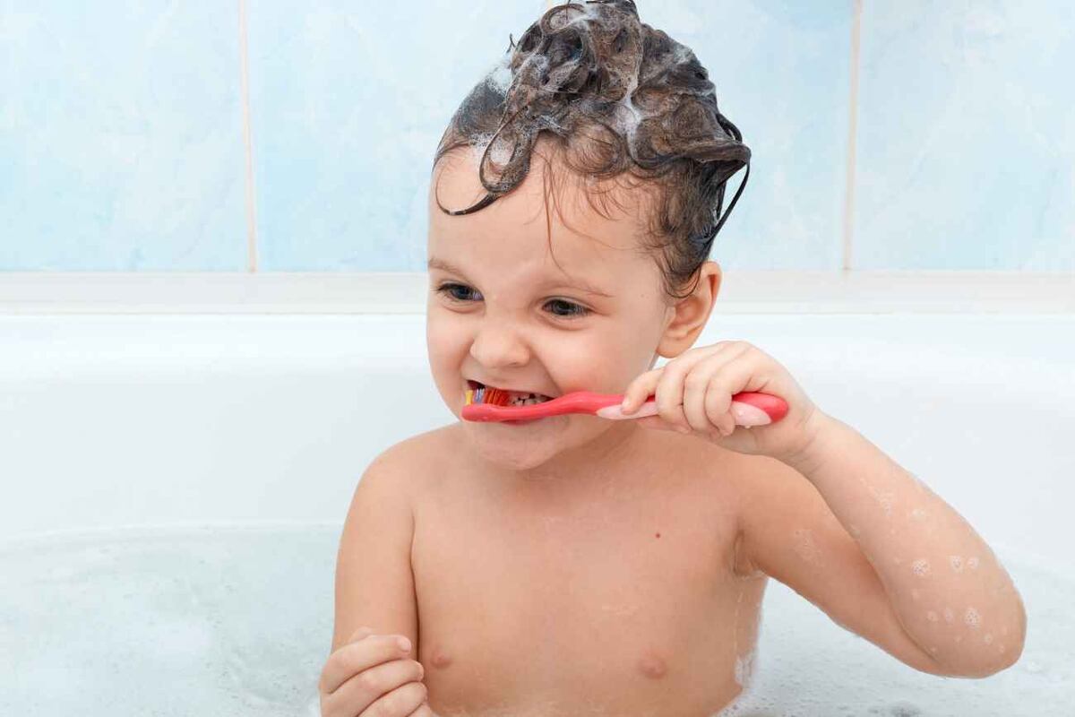 Adorable little baby girl brushing her teeth, taking bath, playing with foam bubbles. Charming little kid glad to be photographed isolated on blue wall in bathroom. Hygiene, child skin and teeth care.